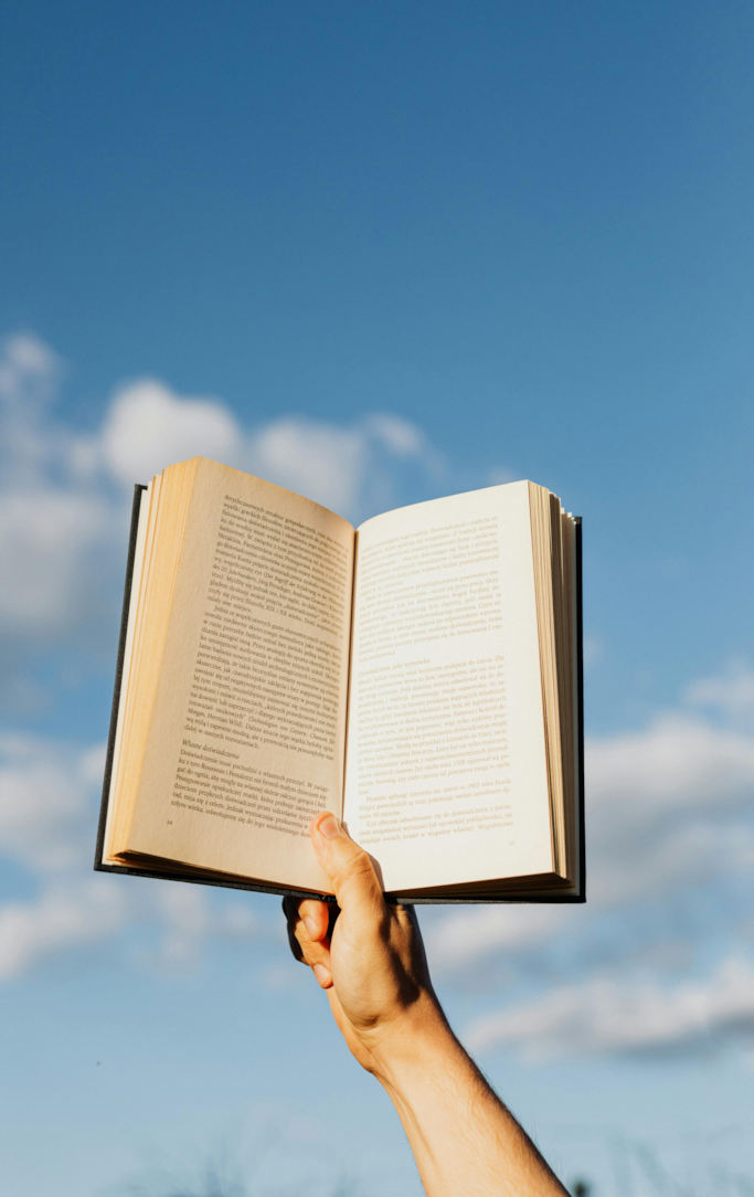 A hand holding an open book aloft against a blue sky with clouds